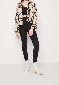 Femme portant une veste courte à motifs beiges, un crop top noir, un legging noir taille haute et des bottines épaisses crème.
