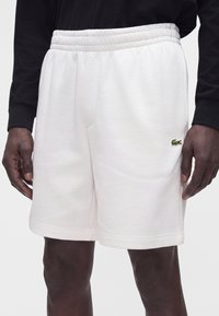 Short blanc à taille élastique avec un petit logo crocodile vert sur la cuisse gauche, porté par une personne à la peau foncée et un top noir à manches longues.