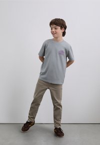 T-shirt gris en coton avec un imprimé graphique violet sur le côté gauche de la poitrine, associé à un pantalon beige clair et des baskets marron avec des accents jaunes.