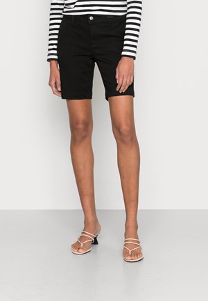 Schwarze Denim-Shorts mit mittelhohem Bund, umgeschlagenem Saum und einer vorderen Knopfverschluss. Kombiniert mit beige Strapsandalen und einem gestreiften Langarmshirt.