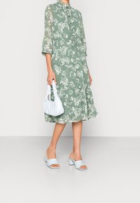 Vestido midi verde con estampado floral, con botones en la parte delantera, dobladillo con volantes y mangas tres cuartos. Combinado con sandalias de tacón bajo en azul claro y un pequeño bolso blanco.