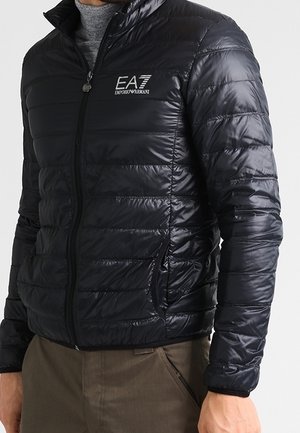 Homme portant une veste matelassée noire avec le logo EA7 Emporio Armani sur un pull à col roulé gris et un pantalon marron.