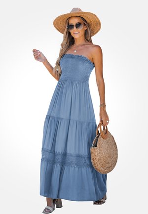 Vrouw draagt een blauwe strapless maxi-jurk, een strooien hoed met brede rand, een zonnebril, houdt een ronde gevlochten handtas vast en draagt sandalen met blokhakken.