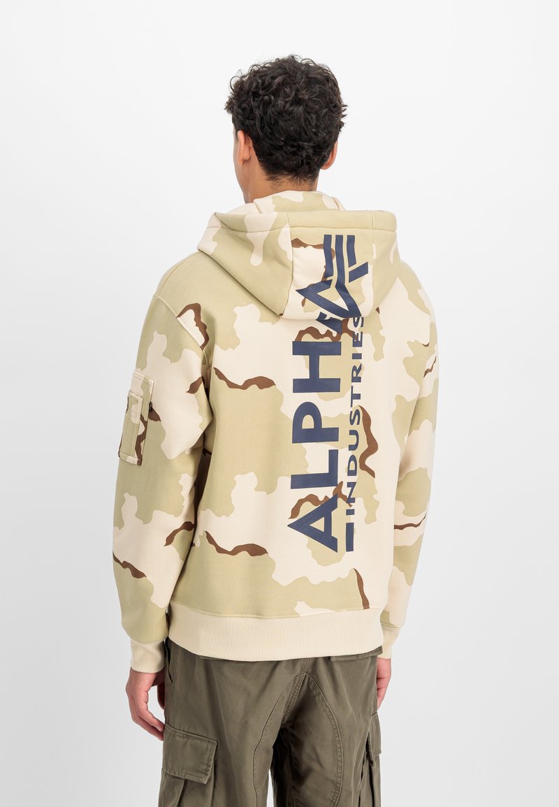 Hoodie camouflage en beige avec des motifs marron, arborant un grand logo "ALPHA INDUSTRIES" en bleu marine au dos et une capuche à cordon.