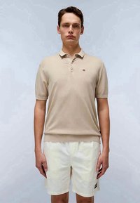 Beige short-sleeve polo shirt with a collar, featuring a small logo on the chest, paired with light-colored shorts. Smooth texture.