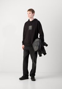 Felpa nera con un patch del logo, abbinata a pantaloni in denim grigio scuro e scarpe da ginnastica nere. Una giacca di pelle nera è appoggiata sul braccio.