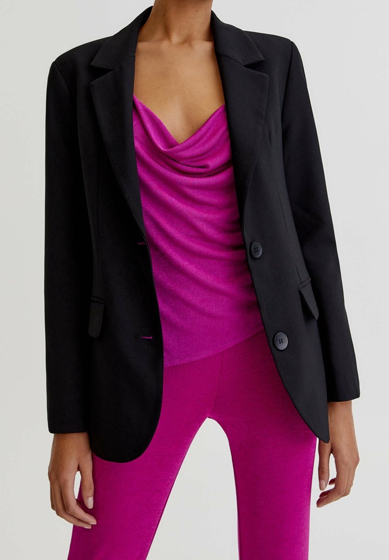 Femme portant un blazer noir cintré sur un top fuchsia drapé et un pantalon fuchsia ajusté assorti, debout devant un fond clair uni.