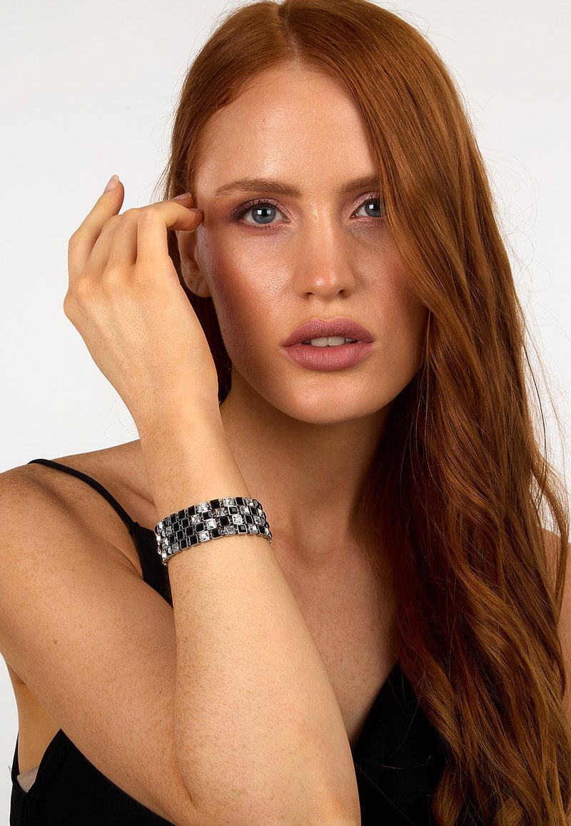 Bracciale nero e argento realizzato con cristalli quadrati, caratterizzato da un pattern a griglia. Indossato al polso di un modello, mette in evidenza il design intricato.