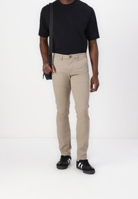 Pantalons en coton beige à coupe slim, avec une fermeture à bouton, des passants de ceinture et des poches latérales, associés à un t-shirt noir à col rond et des chaussures noires.