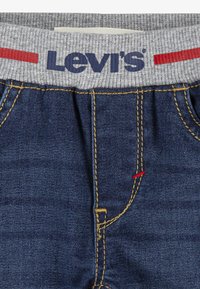 Pantalon en denim avec une ceinture élastique grise ornée du mot « Levi's » en bleu et un accent rouge. Couture bleue et détail rouge à l'avant.