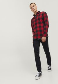 Chemise en flanelle à carreaux rouges et noirs, à fermeture à boutons, en tissu léger, associée à un pantalon slim-fit bleu marine et des baskets noires avec des lacets blancs.