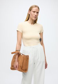 Ljusbrun handväska i mocka med kort handtag, kombinerad med en figursydd ljusbeige t-shirt och lösa vita byxor. Minimalistisk design och neutrala färger.