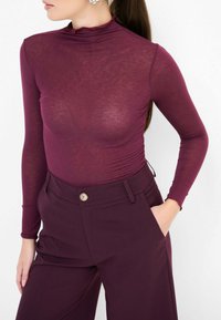 Top bordeaux a maniche lunghe con colletto alto, realizzato in tessuto trasparente. Abbinato a pantaloni tapered a vita alta con dettaglio dei bottoni.