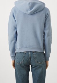 Sudadera con capucha de color azul claro, con puños y dobladillo acanalados, combinada con jeans azules. La tela parece suave con un acabado texturizado.