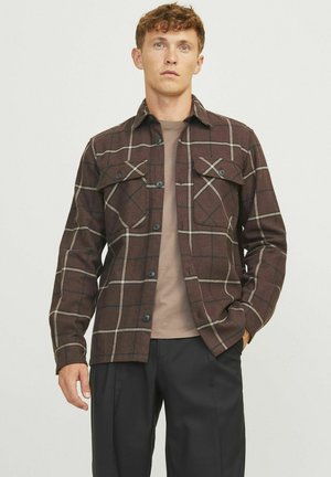 Bruine geruite flanellen shirt met twee borstzakken, zwarte knopen en contrasterende rasterlijnen. Draag over een beige T-shirt en zwarte broek.