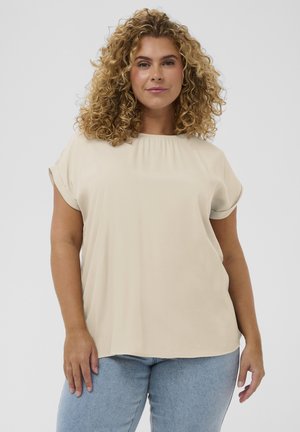 Femme aux cheveux bouclés portant un chemisier beige à manches courtes et un jean bleu clair, debout devant un fond clair uni.