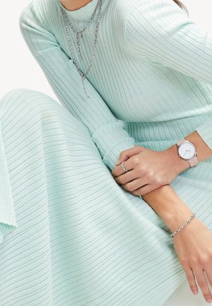 Femme portant une robe en maille côtelée vert menthe clair, un collier chaîne argenté, des bracelets argentés, une bague argentée et une montre avec un bracelet rose pâle.