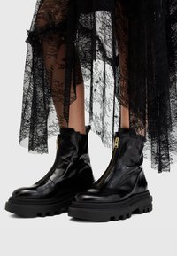 Botas de piel negra hasta el tobillo con suela gruesa, con cierre de cremallera lateral y textura suave, combinadas con una falda de encaje negro transparente.