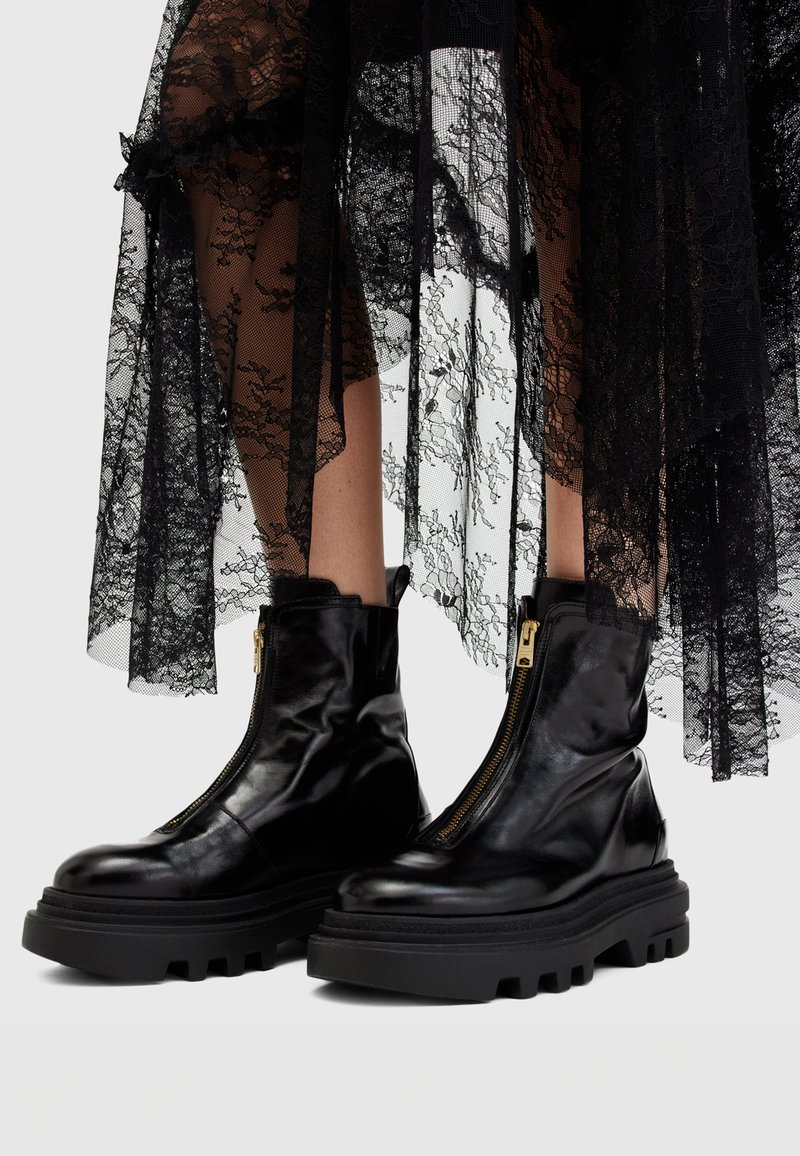 Botas de piel negra hasta el tobillo con suela gruesa, con cierre de cremallera lateral y textura suave, combinadas con una falda de encaje negro transparente.