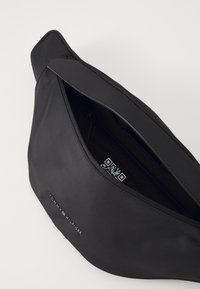 Banane en nylon noir avec une texture lisse, dotée d'un compartiment zippé et d'un logo en relief sur l'extérieur. Code QR visible à l'intérieur.
