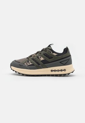 Scarpa da trail verde e nera con una parte superiore testurizzata, tre bande laterali e una suola beige con tappi prominenti e dettagli di branding.