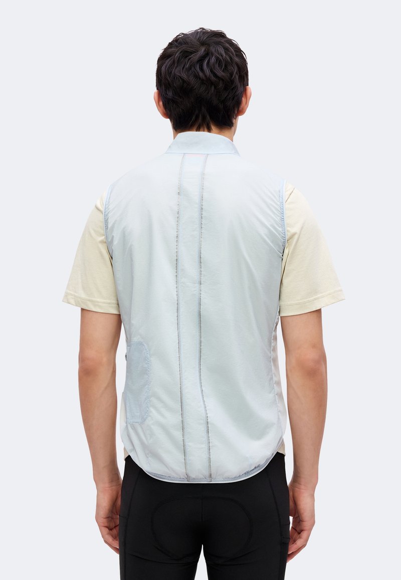 Hombre con chaleco sin mangas azul claro sobre camisa beige de manga corta, visto desde atrás contra un fondo claro y liso.