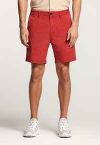 Pantalones cortos de algodón rojos con corte recto, cierre de botón y cremallera, y bolsillos laterales. Usados con zapatillas deportivas blancas.
