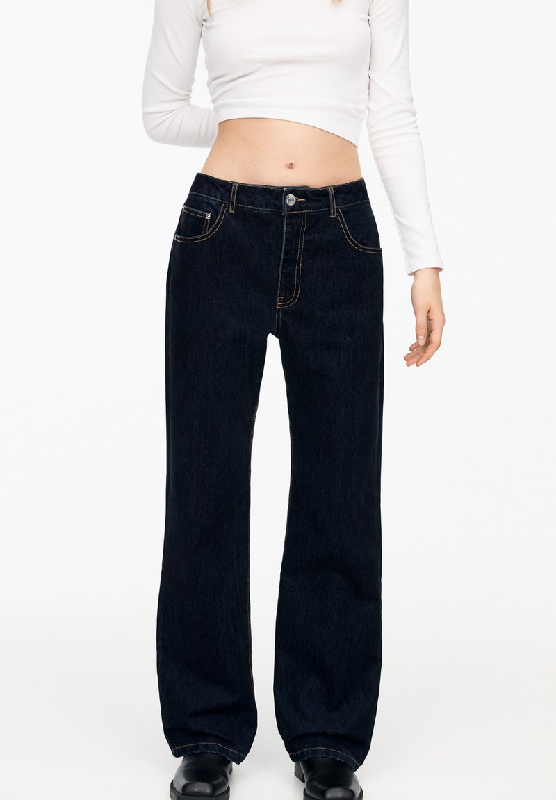 Frau trägt hochtaillierte dunkelblaue Straight-Leg-Jeans, ein kurzes weißes Langarmoberteil und schwarze Stiefel vor einem weißen Hintergrund.