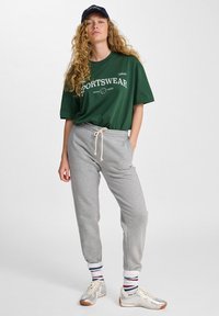 Zielona koszulka z napisem "SPORTSWEAR", szare dresowe spodnie z troczkiem, w paski skarpety i srebrne trampki. Luźny sportowy strój.