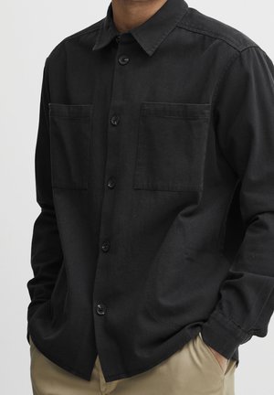 Homme portant une chemise noire à boutons avec deux poches poitrine et un pantalon beige, mains dans les poches, sur fond uni.