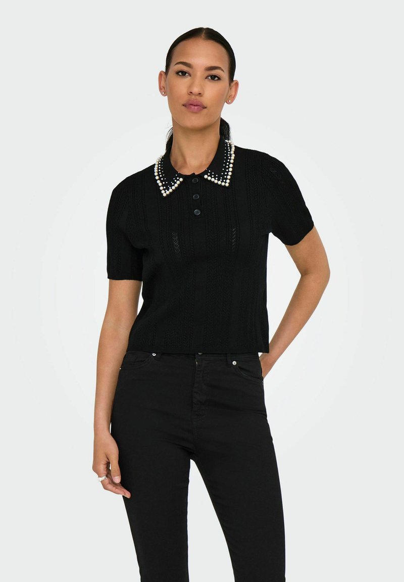 Černý pletený polo top s krátkým rukávem, zdobený límcem s perlovými akcenty, zapínáním na knoflíky a texturovaným vzorem. Nosený s černými kalhotami.