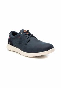 Zapatos sintéticos azul marino con una textura suave, diseño minimalista, cordones y una suela de goma blanca. Presenta un acento marrón en la lengüeta.