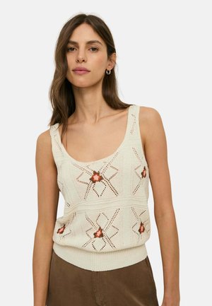 Mujer con cabello castaño que lleva un top sin mangas de punto crema con patrones florales y pantalones marrones sobre un fondo liso.