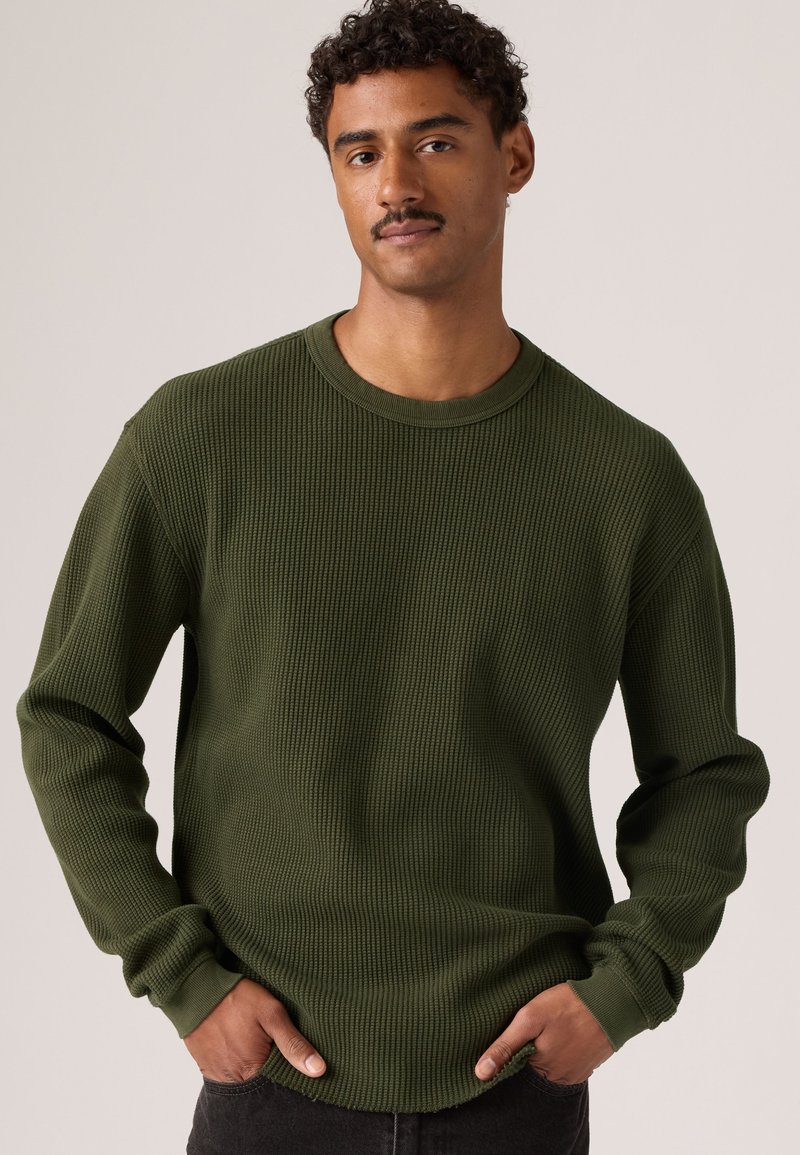 Homme aux cheveux courts et bouclés avec une moustache portant un pull vert texturé à manches longues, les mains dans les poches, debout devant un fond uni.