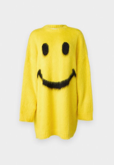 Gul strikket sweater lavet af fluffy stof med et sort smiley-ansigt design, der har store øjne og et bredt smil.