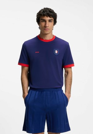 Mann trägt ein marineblaues Kurzarmhemd mit rotem Kragen und Manschetten, der Aufschrift „BOSS“ und passenden marineblauen plissierten Shorts, Hände in den Taschen.