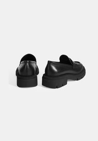 Zwarte leren loafers met een chunky zool, met een ronde neus en een minimalistisch ontwerp. De achterkant toont een gladde textuur en subtiele details.