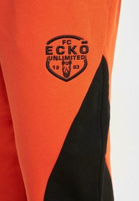 Orange joggers featuring black side panels and an embroidered logo with a shield shape, text, and a stylized animal head.