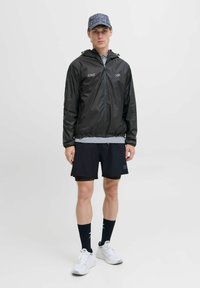 Czarna kurtka przeciwwiatrowa z kapturem, szary t-shirt pod spodem, czarne szorty, białe trampki oraz niebieska czapka w wzory. Skarpetki są ciemne z wzorem.