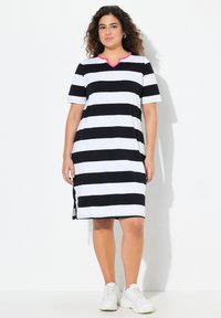 Schwarz-weiß gestreiftes Kleid mit V-Ausschnitt und pinken Akzenten, kurzen Ärmeln und Seitenschlitzen. Getragen mit weißen Sneakers und entspannter Passform.