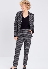 Sort-hvid ternet blazer og matchende bukser med en afslappet pasform. Båret over en sort V-hals sweater. Glat stof med definerede linjer.