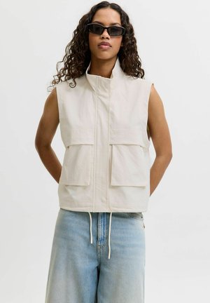 Femme aux cheveux bouclés portant des lunettes de soleil noires, un gilet beige sans manches avec de grandes poches et un jean bleu clair sur un fond uni.