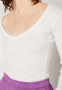 Balta, raukšlėta ilgomis rankovėmis palaidinė su V formos dekoltė, derinama su aukštai supjaustyta purpurinio spalvos elastinga juosmens sijonu. Minkšta tekstūra ir tvirtas prigludimas.
