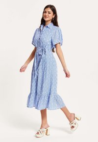 Blue, knee-length dress with puffed sleeves, button front, and a tie waist. Features a tiered hem and floral pattern, paired with chunky heels.
