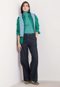Camicetta verde, dolcevita a righe, maglione blu appoggiato sulle spalle, pantaloni in denim scuro, sandali verdi e beige, tessuto a trama.