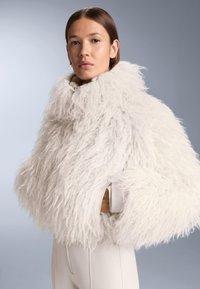 Kobieta o prostych brązowych włosach ma na sobie puszystą, białą kurtkę o obszernym kroju z wysokim kołnierzem oraz białe dopasowane spodnie na szarym tle.