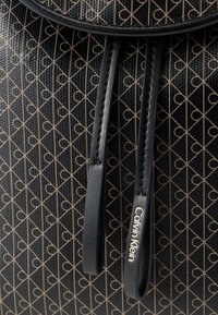 Μαύρη τσάντα με υφή και επαναλαμβανόμενο μπεζ γεωμετρικό σχέδιο, με δύο μαύρα λουριά, ένα λουρί με ανάγλυφο λογότυπο Calvin Klein σε χρυσά γράμματα.