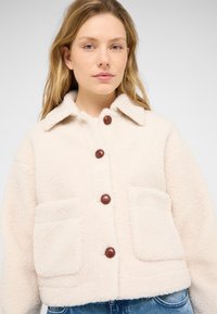 Fleece-Jacke in Cremefarbe mit kragenlosem Hals, braunen Knopfverschluss und zwei aufgesetzten Taschen auf der Vorderseite. Lockeres und strukturiertes Design.