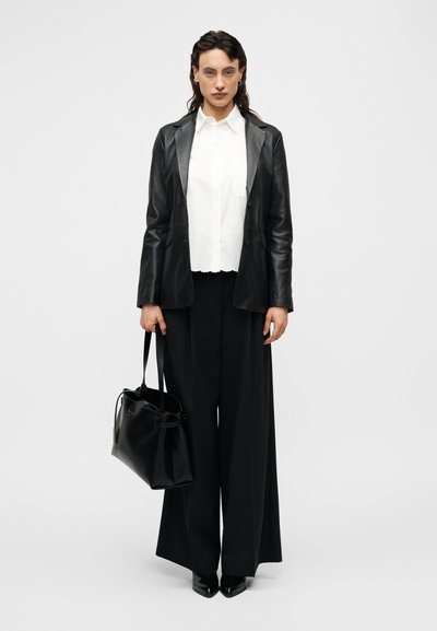 Μαύρο δερμάτινο σακάκι πάνω από λευκό πουκάμισο με γιακά, σετ με φαρδιά μαύρη παντελόνα. Κρατείται μαύρη δερμάτινη τσάντα σε ένα χέρι.