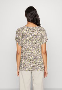 Mujer vestida con una blusa de mangas cortas de estampado floral con pequeñas flores moradas y amarillas, combinada con pantalones beis claros, vista desde atrás.